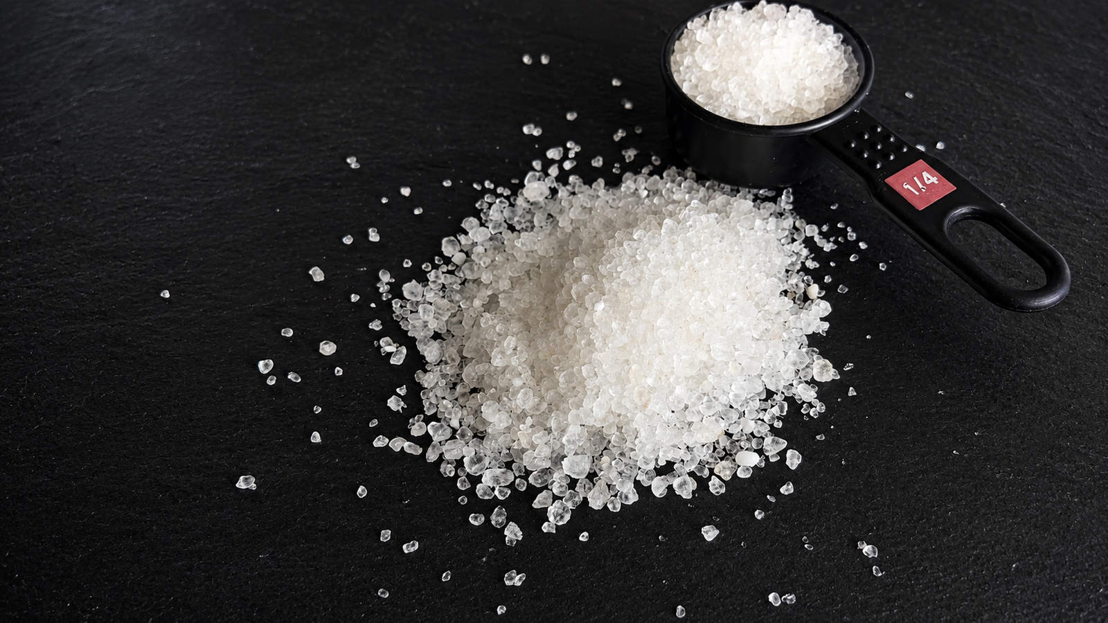 Magnesium chloride crystals beside a measuring scoop on a dark surface.